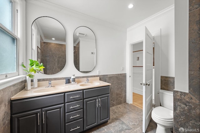 full bath featuring tile walls, crown molding, toilet, double vanity, and a wainscoted wall