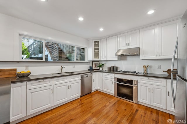 kitchen with under cabinet range hood, dark countertops, a sink, appliances with stainless steel finishes, and white cabinetry