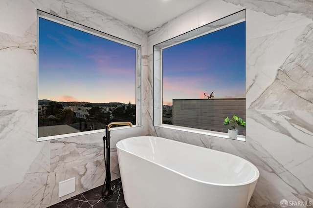 full bath with marble finish floor, stone wall, and a freestanding tub