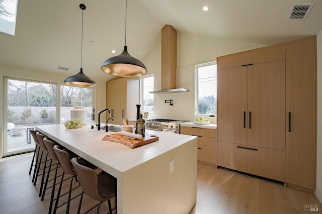 kitchen with wall chimney exhaust hood, hanging light fixtures, light brown cabinets, double oven range, and an island with sink