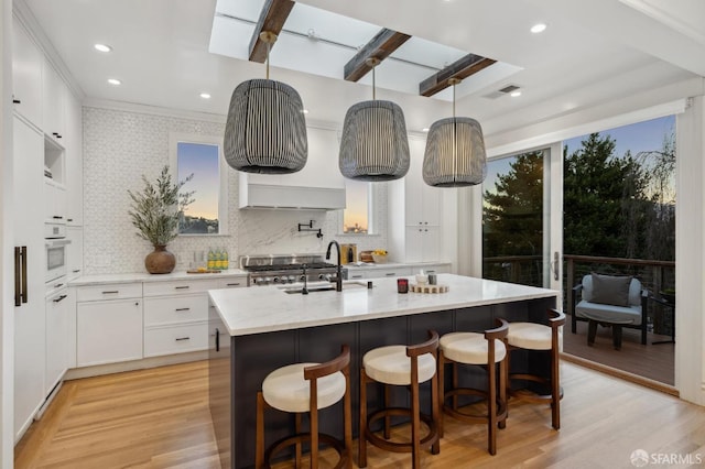 kitchen featuring a skylight, white cabinetry, an island with sink, sink, and light hardwood / wood-style floors