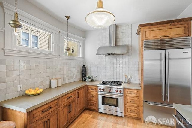 kitchen featuring wall chimney exhaust hood, light countertops, high end appliances, light wood finished floors, and brown cabinetry