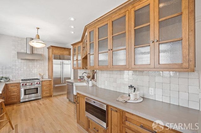 kitchen with brown cabinetry, built in appliances, wall chimney exhaust hood, and light wood finished floors
