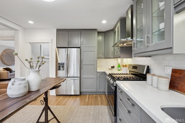 kitchen featuring wall chimney range hood, light hardwood / wood-style flooring, appliances with stainless steel finishes, tasteful backsplash, and light stone counters