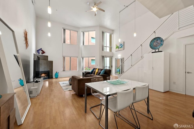 dining space featuring a ceiling fan, a towering ceiling, light wood-type flooring, stairway, and a warm lit fireplace