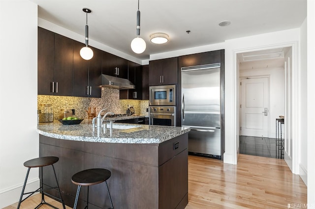 kitchen with built in appliances, kitchen peninsula, dark brown cabinets, light stone counters, and light hardwood / wood-style floors