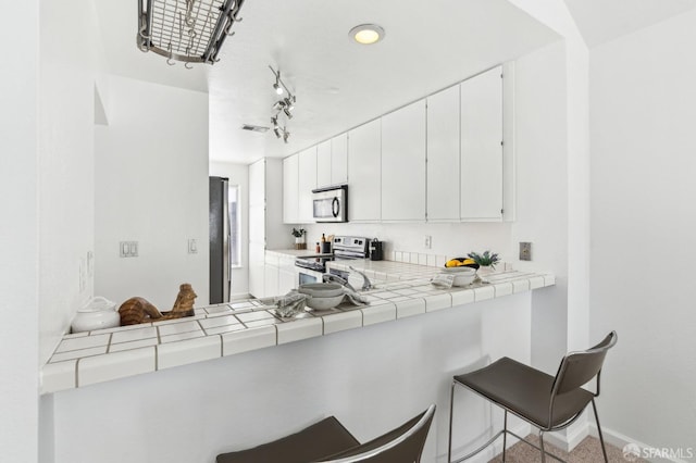 kitchen featuring kitchen peninsula, tile counters, stainless steel appliances, a breakfast bar, and white cabinetry