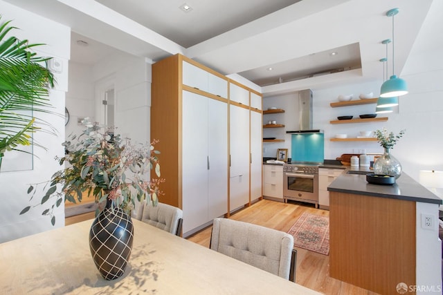 kitchen with a tray ceiling, open shelves, light wood-style flooring, wall oven, and wall chimney range hood