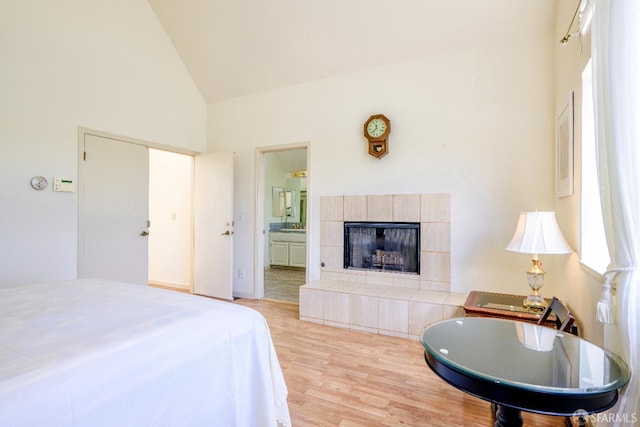 bedroom featuring high vaulted ceiling, wood finished floors, a tiled fireplace, and ensuite bath