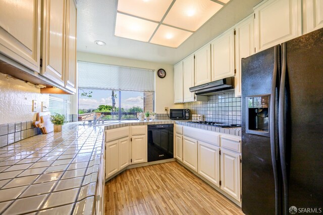 kitchen with black appliances, tile countertops, under cabinet range hood, tasteful backsplash, and white cabinetry