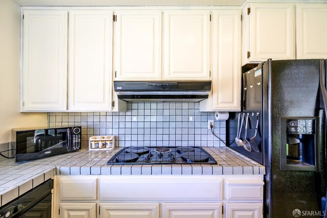 kitchen with tile counters, black appliances, under cabinet range hood, white cabinetry, and backsplash