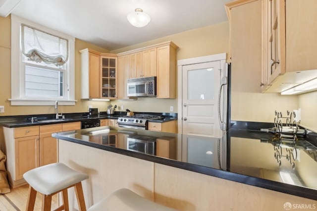 kitchen with a breakfast bar area, stainless steel appliances, glass insert cabinets, light brown cabinets, and a sink