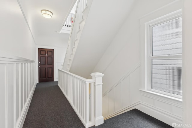 hallway featuring wainscoting, dark carpet, and a decorative wall