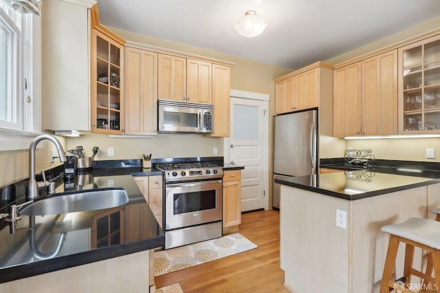 kitchen with dark countertops, light brown cabinetry, appliances with stainless steel finishes, a sink, and light wood-type flooring