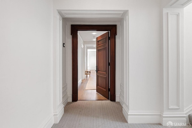 corridor featuring a decorative wall, light colored carpet, and baseboards