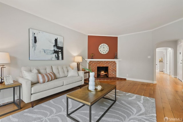 living area featuring arched walkways, hardwood / wood-style flooring, crown molding, a brick fireplace, and baseboards