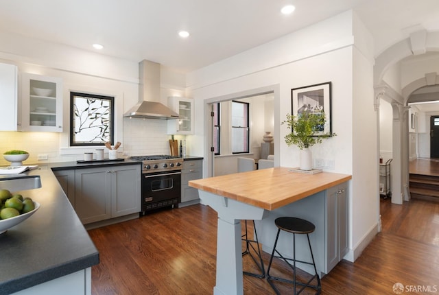 kitchen featuring high end stove, wall chimney range hood, gray cabinets, dark hardwood / wood-style floors, and butcher block counters