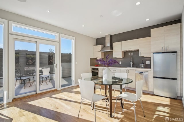 kitchen featuring appliances with stainless steel finishes, tasteful backsplash, sink, wall chimney range hood, and light hardwood / wood-style flooring