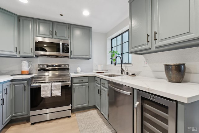kitchen with sink, gray cabinets, appliances with stainless steel finishes, light stone counters, and beverage cooler
