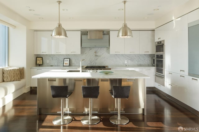 kitchen with white cabinetry, decorative light fixtures, an island with sink, and wall chimney range hood