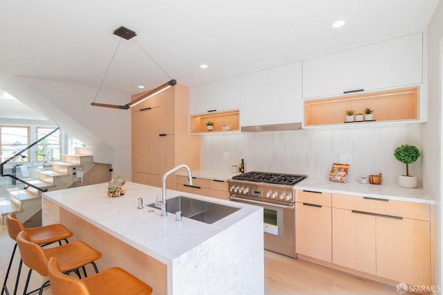 kitchen with light brown cabinetry, an island with sink, sink, hanging light fixtures, and stainless steel range