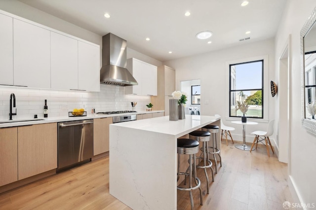 kitchen with stainless steel dishwasher, wall chimney exhaust hood, sink, white cabinetry, and a kitchen island