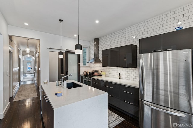 kitchen featuring a center island with sink, hanging light fixtures, wall chimney exhaust hood, a barn door, and appliances with stainless steel finishes