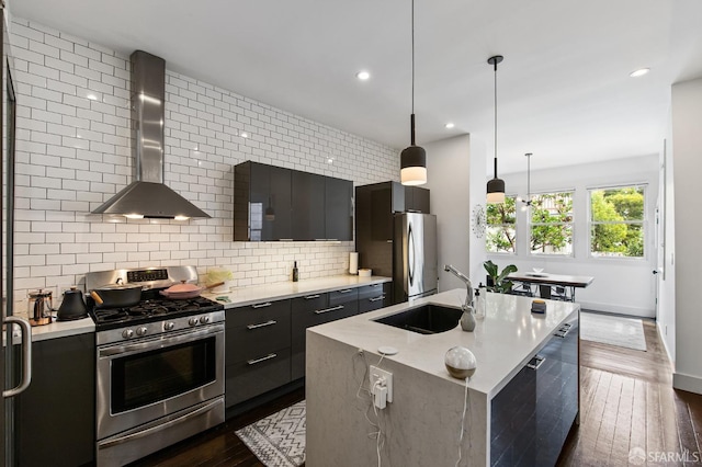 kitchen featuring sink, wall chimney exhaust hood, an island with sink, decorative light fixtures, and stainless steel appliances