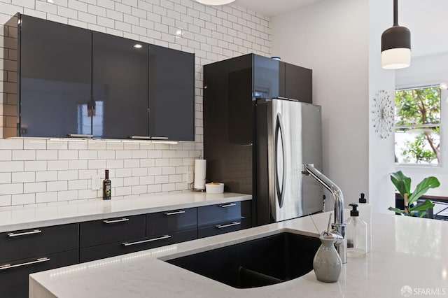 kitchen featuring sink, light stone counters, backsplash, stainless steel fridge, and pendant lighting