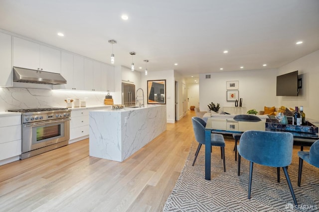 kitchen with pendant lighting, high end stove, a kitchen island with sink, white cabinets, and light hardwood / wood-style flooring