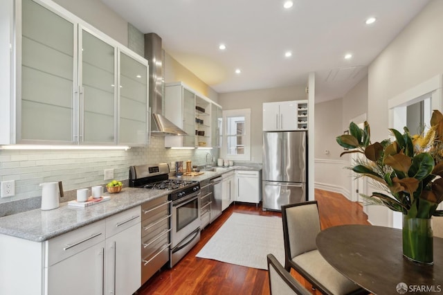 kitchen with wall chimney exhaust hood, glass insert cabinets, light stone counters, stainless steel appliances, and white cabinetry