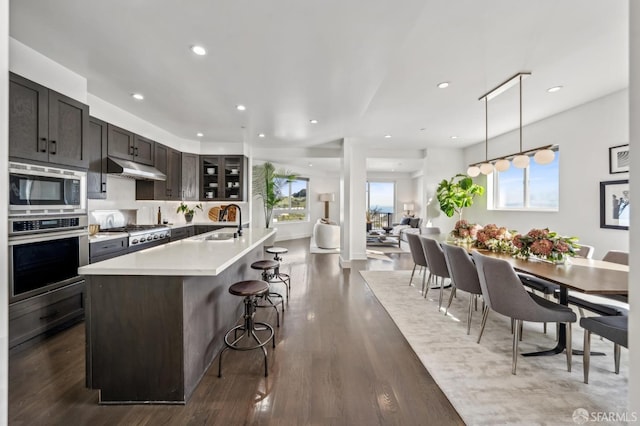 kitchen featuring a sink, light countertops, under cabinet range hood, appliances with stainless steel finishes, and a kitchen bar