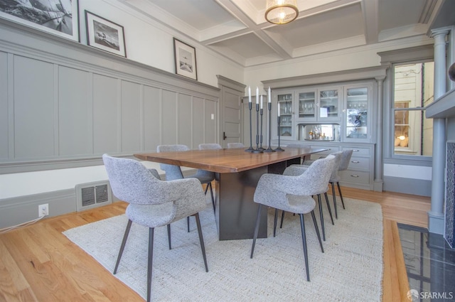 dining area with light wood finished floors, a decorative wall, coffered ceiling, beam ceiling, and crown molding