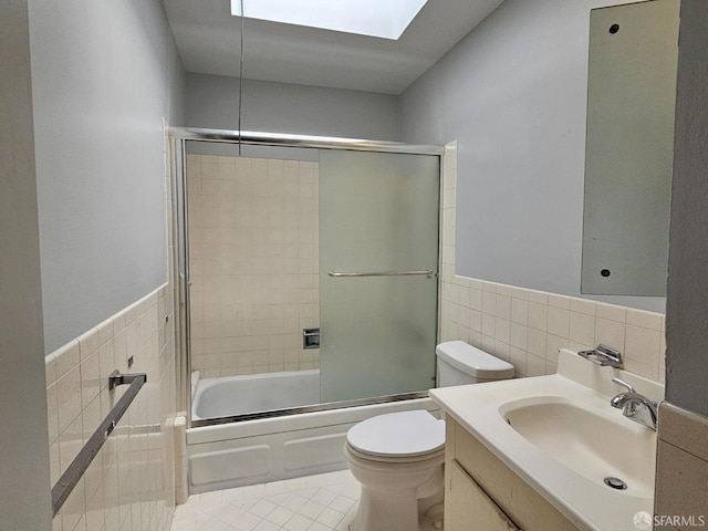 full bathroom featuring a skylight, bath / shower combo with glass door, vanity, tile walls, and toilet