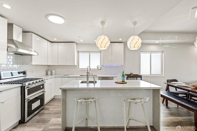 kitchen with light wood finished floors, range with two ovens, wall chimney exhaust hood, a sink, and backsplash