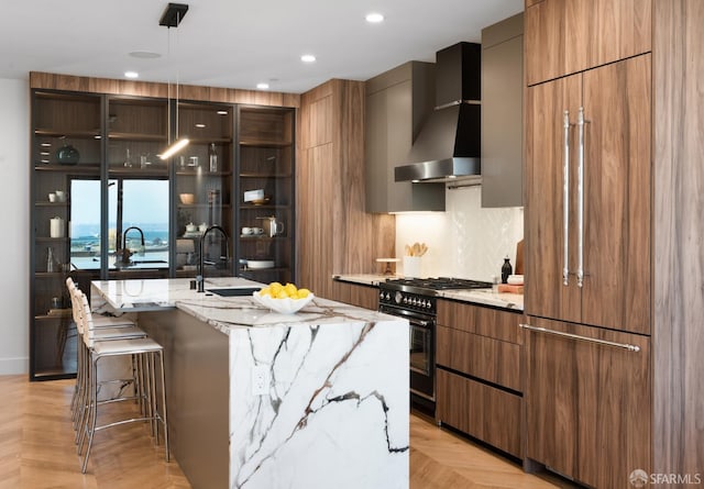 kitchen with a kitchen island with sink, wall chimney exhaust hood, high quality appliances, light stone counters, and light parquet flooring