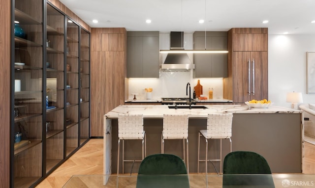 kitchen with a kitchen breakfast bar, light parquet floors, a kitchen island with sink, light stone countertops, and wall chimney exhaust hood