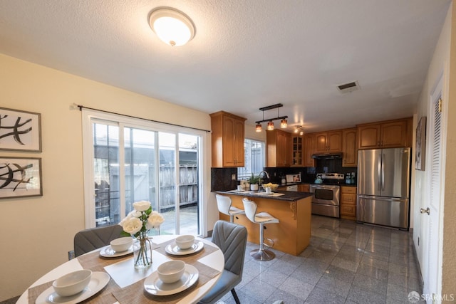 kitchen featuring dark countertops, glass insert cabinets, under cabinet range hood, appliances with stainless steel finishes, and a peninsula