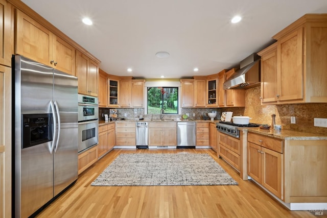 kitchen with light stone countertops, glass insert cabinets, light wood-style flooring, appliances with stainless steel finishes, and wall chimney exhaust hood