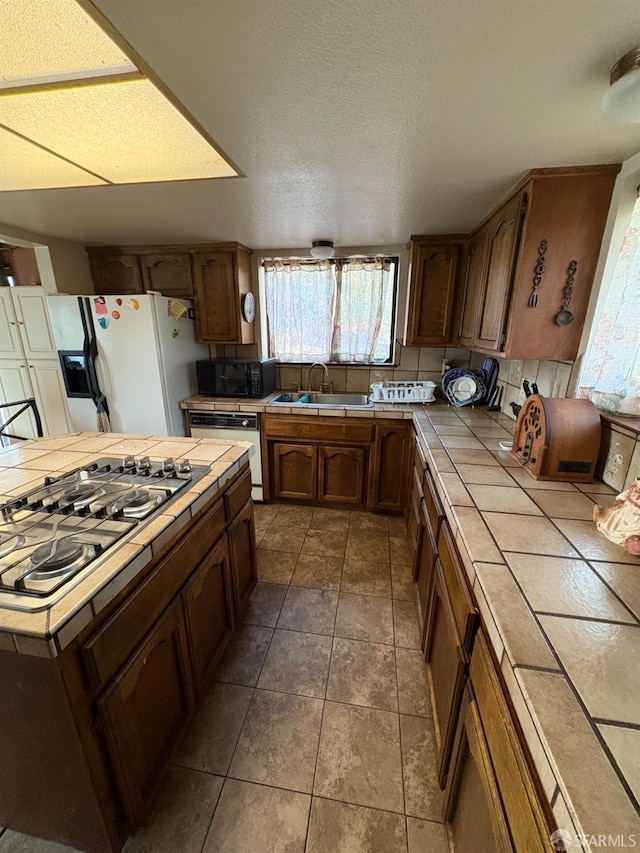 kitchen with sink, dishwasher, tile countertops, stainless steel gas stovetop, and white refrigerator with ice dispenser