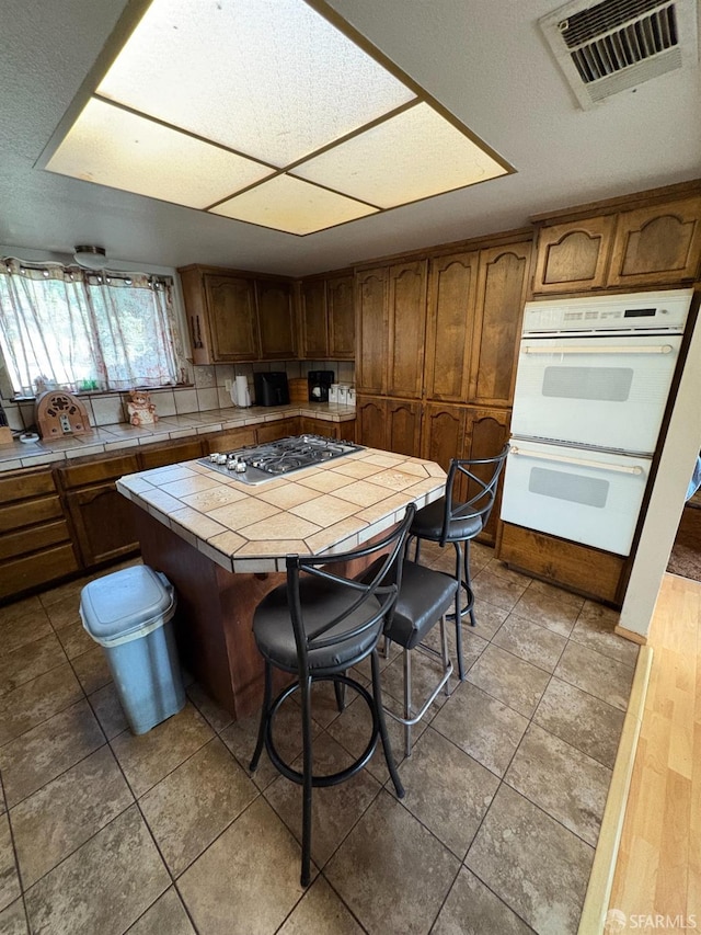 kitchen with tile countertops, tile patterned flooring, a kitchen breakfast bar, stainless steel gas stovetop, and white double oven