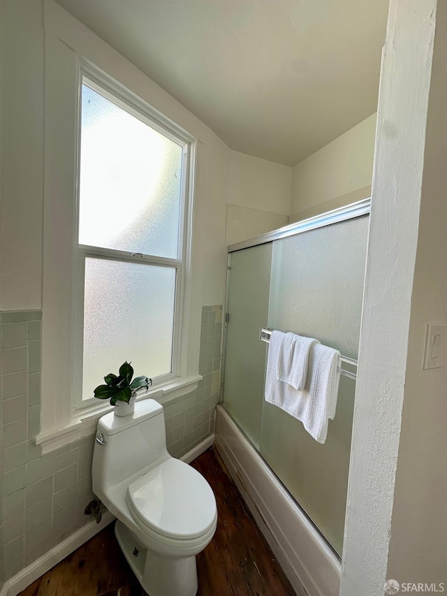 bathroom with tile walls, wood-type flooring, combined bath / shower with glass door, and toilet