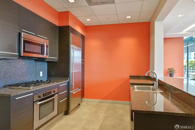 kitchen featuring backsplash, dark stone countertops, sink, a paneled ceiling, and appliances with stainless steel finishes