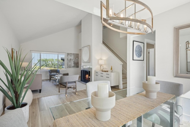 dining space with high vaulted ceiling, a lit fireplace, stairs, light wood-type flooring, and a chandelier