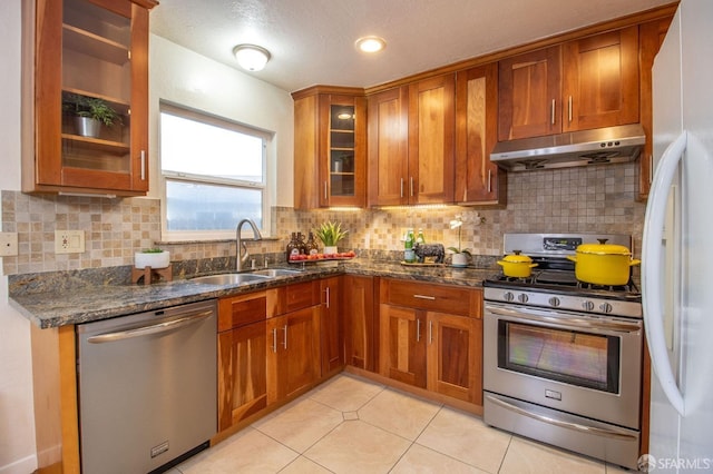 kitchen featuring brown cabinetry, dark stone counters, a sink, stainless steel appliances, and under cabinet range hood