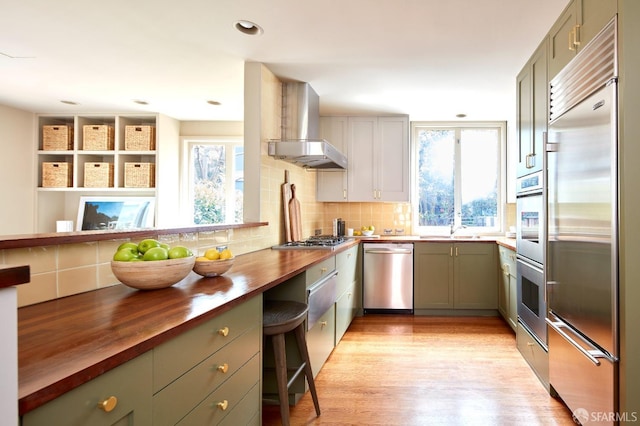 kitchen featuring butcher block counters, tasteful backsplash, wall chimney range hood, and stainless steel appliances