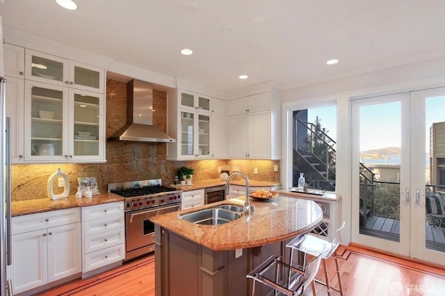 kitchen with light stone counters, a kitchen island with sink, sink, wall chimney range hood, and stainless steel stove