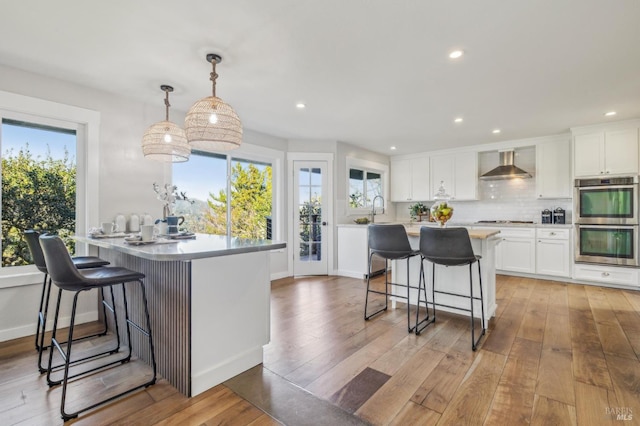 kitchen featuring decorative light fixtures, white cabinets, stainless steel double oven, light hardwood / wood-style floors, and wall chimney range hood