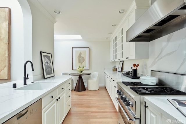 kitchen featuring wall chimney range hood, stainless steel appliances, a sink, light stone countertops, and light wood finished floors