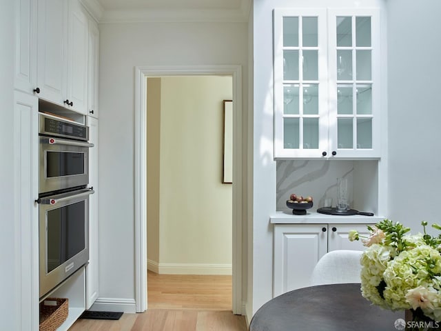 kitchen with double oven, light wood-style floors, white cabinetry, baseboards, and ornamental molding